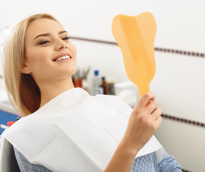 A woman sitting in a dental chair, examining her teeth with a dental mirror and feeling happy with the results of her cosmetic dental treatment in the clinic.