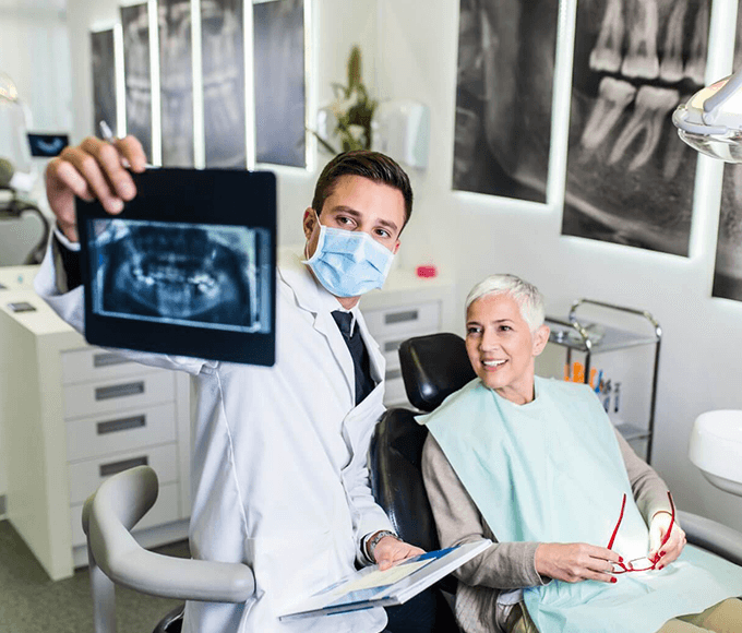 A male dentist in a white coat and surgical mask confidently presents a dental x-ray to a smiling female patient with short grey hair, who is seated in the dental chair. The modern dental clinic is well-equipped, with bright lights and multiple dental x-ray images displayed in the background, illustrating a professional consultation.