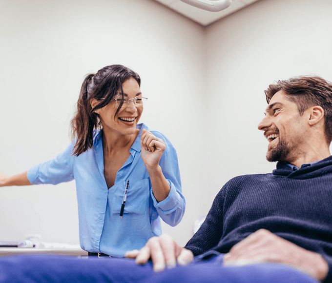 A female dentist in a light blue shirt and protective eyewear is pointing at a dental x-ray on a monitor, sharing a light-hearted moment with a male patient. The patient, seated in the dental chair, looks on with a relaxed and cheerful expression, reflecting a positive dentist-patient interaction.