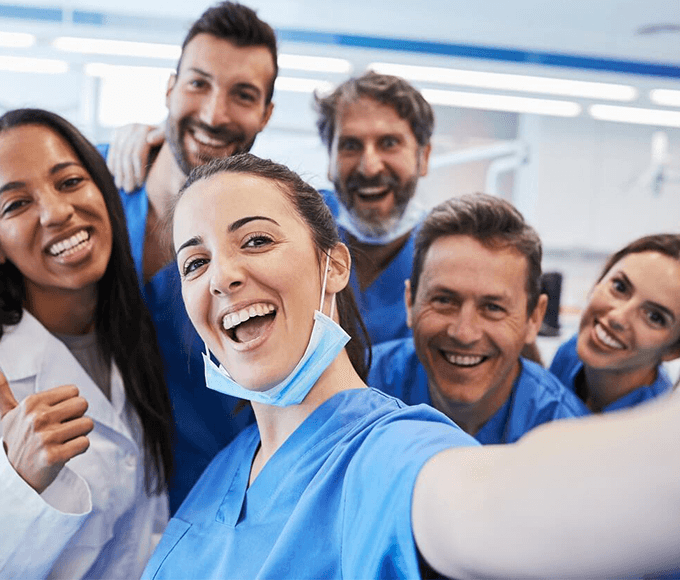 A cheerful dental team composed of six diverse professionals taking a group selfie, with warm smiles and positive demeanors in a dental clinic environment. They wear blue scrubs and have their surgical masks pulled down, emphasizing a friendly and professional dental care team.