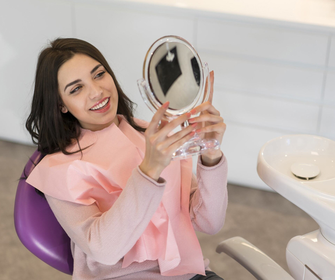 A woman in a dentist's office, sitting in a chair and examining her straight smile in a mirror after having her braces removed, looking happy and satisfied with the results.