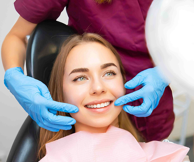 A young woman smiles happily while looking into a hand mirror after a teeth whitening procedure at the dentist's office. The dentist, wearing blue gloves, gently touches her cheeks to showcase the results.