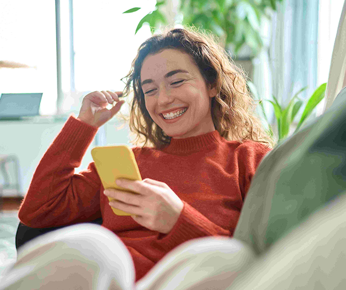 A woman wearing a white blouse is sitting at a desk, smiling and looking to the side. She is holding a pen and appears to be engaged in a conversation or a video call. A laptop and a coffee mug are visible in the foreground, and a plant is in the background.